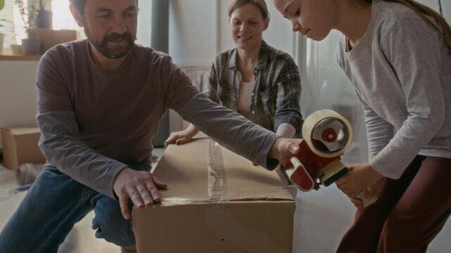 Young couple packing cardboard boxes with fragile objects and other household stuff while their little daughter helping them seal packages with adhesive tape, then family giving each other high five