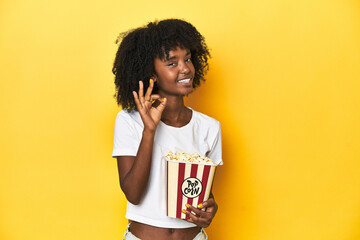 Teen girl with popcorn, cinema concept on yellow backdrop cheerful and confident showing ok gesture.