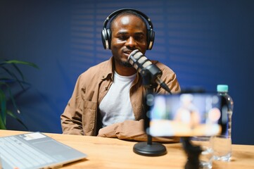 Black vlogger filming podcast on smartphone in studio. Cheerful blogger