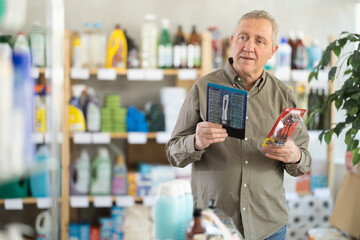 Elderly man is standing in a supermarket and holding razors from different manufacturers in hands,...