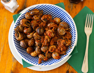 On wooden table there is plate filled to brim with boiled snails. Shellfish snail cooked with minced meat and tomato dressing. Chopsticks are served with food.