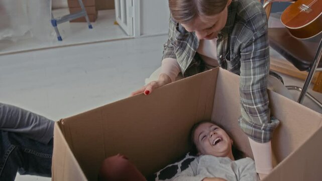Cheerful little girl lying in cardboard box while her parents sitting on the floor of domestic room during relocation, tickling her and playing with their daughter