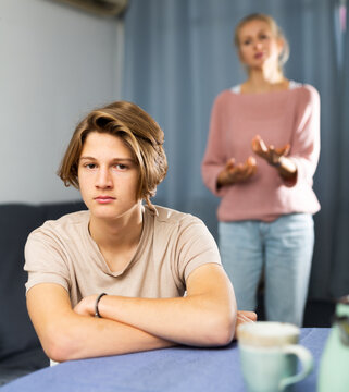 Frowning teenager with bored expression listening to reprimanding from his mother at home..