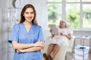 Obraz premium Smiling young lady wearing medical scrub standing at treatment room in front of female patient lying at examination couch.