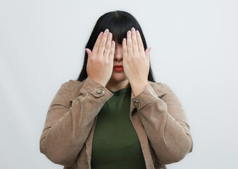 Woman covering her eyes with both hands, wearing a green shirt and brown jacket. Symbolizing...