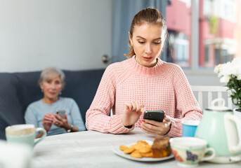 Young woman sitting at table at home and using her smartphone. Her senior grandmother sitting on sofa in background and using smartphone too.