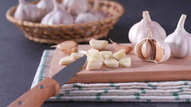 Sliced garlic cloves on a cutting board on a black stone table. Chopped garlic on a cutting board, in background a pile of garlic unpeeled.