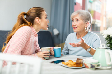 Caucasian young woman and her mature mother sitting at table, talking and drinking tea.
