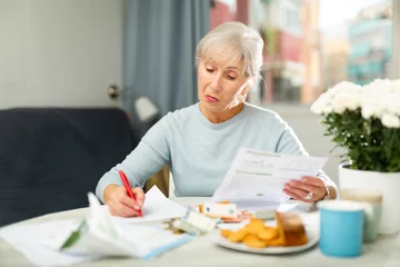 Fotobehang Grey haired thoughtful woman sitting at table and analysing financial documents. She's making notes on paper and counting money. © JackF