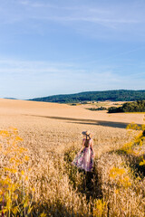 A woman in a flowing floral dress and a wide-brimmed hat stands in a golden wheat field under a clear blue sky. The warm sunlight enhances the peaceful and dreamy atmosphere.