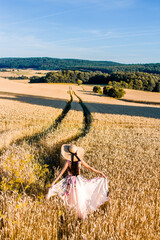 A woman in a flowing floral dress and a wide-brimmed hat stands in a golden wheat field under a...