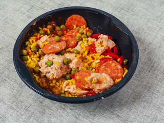 Spanish paella dish in black round plastic tray on light color table cloth. Popular food with rice and sea products and meat. Fast food product.