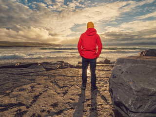 Man in red jacket on a rough stone rock shore looking at stunning sunset sky and ocean in the...