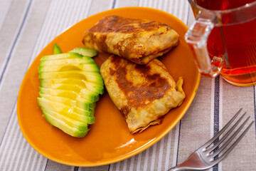 There are pancakes with liver and egg filling on plate, supplemented with half chopped avocado. Cup of hot black tea is served for breakfast.