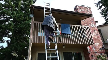 Senior man climbing a shaky aluminum extension ladder with a leaf blower to clean a rooftop of tree debris, property maintenance

