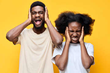 Young African couple expressing frustration with loud noise, covering their ears against a bright yellow background Emotions include anger and annoyance