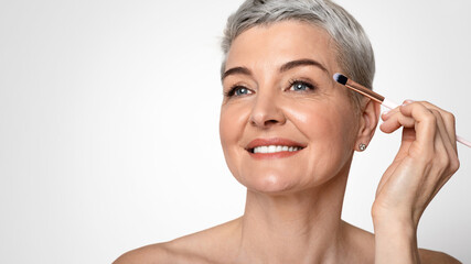 Fototapeta premium Aging Gracefully Concept. Portrait Of Beautiful Mature Woman Applying Eye-Shadows And Looking Away Over Light Studio Background, Closeup