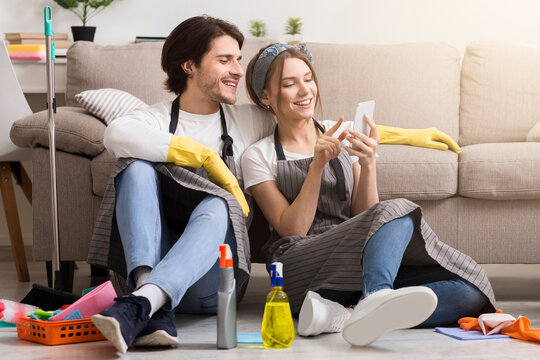 Smiling young couple relaxing with smartphone after spring-clean apartment, sitting on floor with lots of detergents and cleaning supplies, free space