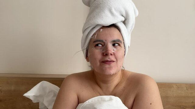 4K video. Young adult white woman with towel on head, bare shoulders is put on cosmetic textile mask on female face in hotel bed room. Concept of Mother Day, rest, relax, skin protection, March 8, spa