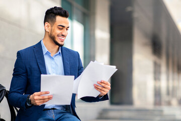 Excited young arab man in formal outfit sitting on the street next to business building, holding documents or CV, having backpack by him, getting ready for job interview, panorama with copy space