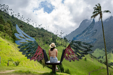 Naklejka premium A woman in casual attire and a straw hat sits on a colorful winged bench overlooking the lush green hills and iconic wax palms of Valle del Cocora in Colombia under a partly cloudy sky.