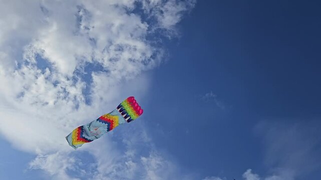 Bottom view of colorful kite with intricate patterns soaring high in the bright blue sky, surrounded by scattered clouds.
