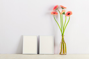 Pink gerbera flowers in a glass vase and two blank frames on a light background. Interior...
