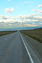 Naklejka premium A two-lane paved road crosses a wide steppe towards a high snow-covered mountain range in the clouds on an early summer morning.