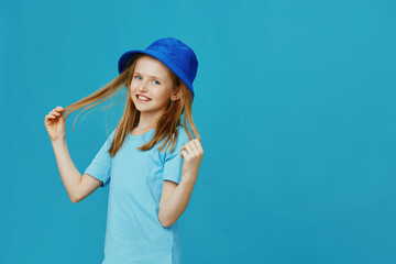 Cheerful girl with long hair in a blue hat, smiling and playing with her hair against a vibrant blue background, perfect for children s fashion themes