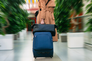 Girl in the brown coat pulls a blue suitcase through an airport. The motion blur effect emphasizes...