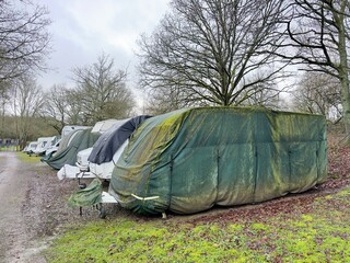 A number of caravans are in an outdoor storage area.They are protected by various covers in various states of condition. Trees in background.
