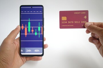 The man's left hand holds a smartphone with a graphic display of stock price movements and his right hand holds a credit card