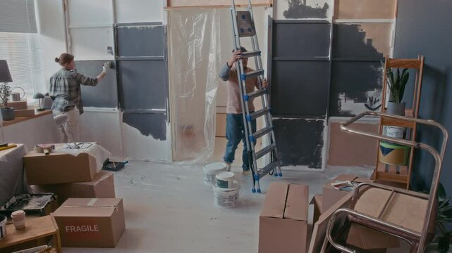 Timelapse of young man and woman doing renovation work in living room of their new apartment painting walls, carrying packed cardboard boxes and repairing household stuff