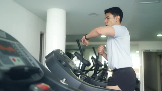 Healthy Asian man checks heart rate and calories burned on smart watch screen at fitness gym. Sportsman sport training workout cardio exercise running on treadmills. Healthcare technology concept.
