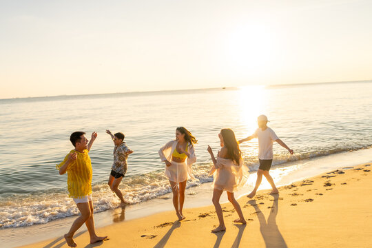 Happy man and woman travel nature ocean on summer beach holiday vacation. Group of Asian people friends enjoy and fun outdoor active lifestyle playing in the sea together at tropical island at sunset.