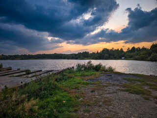 A tranquil lakeside scene at sunset, featuring dramatic clouds and a vibrant sky. the peaceful water reflects the rich hues of the setting sun, while lush greenery surrounds the area, creating a calm