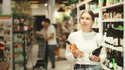 Young woman holds bottles of wine in her hands, reading labels and stands near the shelves with alcohol in a hypermarket against the background of customers. High quality 4k footage