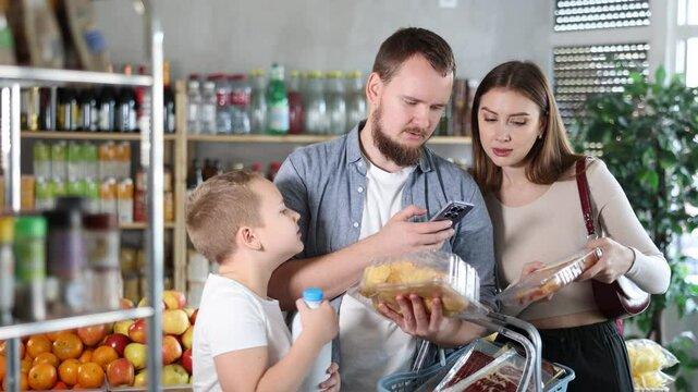 Interested tween boy with friendly young father and mother scanning barcodes on packages with fresh raw cut poultry parts using phone while shopping in supermarket, checking product information and
