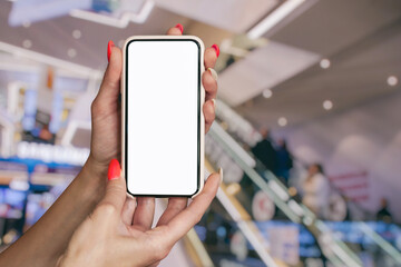 Woman with red manicured nails is holding mockup smartphone with a blank white screen in a shopping mall.