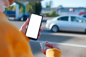 Woman is holding a mockup smartphone with a blank white screen and money at a gas station, showcasing mobile payment and outdoor transactions.