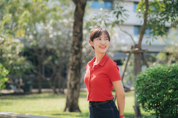 Fototapeta premium Asian cheerful woman in red shirt enjoys sunny day in green park, surrounded by trees and natural light