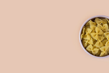 Top view of a porcelain bowl of raw italian durum wheat farfalle pasta against a light ochre...