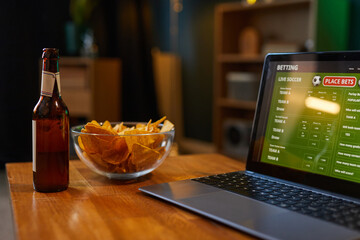 Scene showing laptop displaying live soccer game with snacks and beer on wooden table. Atmosphere from cozy room setting