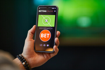 Close-up of hand holding smartphone with soccer betting app interface showing bet options and match information in background. Image depicts technology and sports interaction