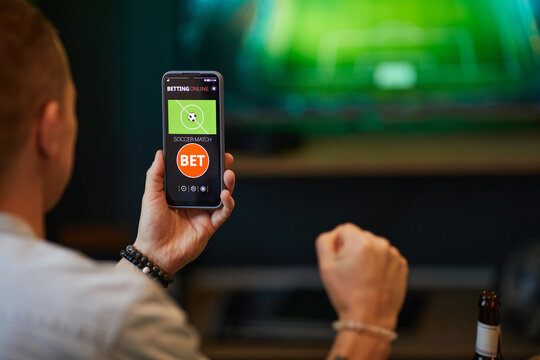 Person engaged in online soccer betting using smartphone while watching game on TV screen. Phone displaying betting interface with options