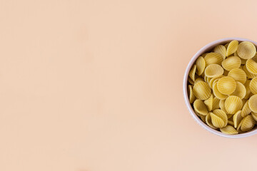 Top view of a porcelain bowl of raw italian durum wheat orecchiette pasta against a light ochre...
