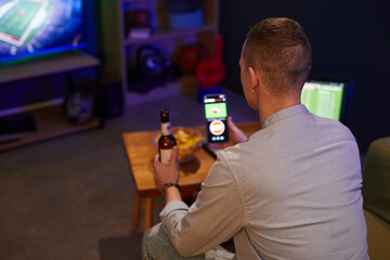 Person seen from behind enjoying a football game on TV while holding a beer and smartphone. Relaxed...