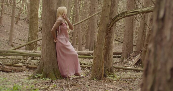 Young Ebony woman, with long blonde hair, wearing a pink gown. She leans against the trunk of a Sweetgum tree, in a forest. She plays with her hair as she looks to, and away from camera, Wide shot.