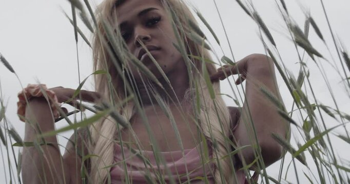 Young ebony woman, with long blonde hair. She is wearing a pink gown, with a matching scrunchie on her right wrist. She runs the tall grass between her fingers, looking at camera. 60fps slow motion.