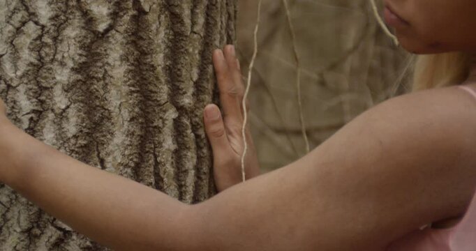 The hands of an ebony girl run along the bark of a sweetgum tree. A pink scrunchie on her right wrist. Right hand runs down along the bark, while left hand goes sideways, away from her. .